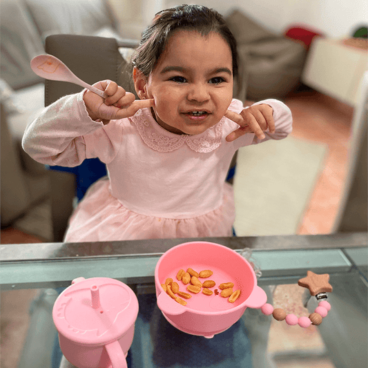 Bebé sentada en la mesa comiendo con su vajilla infantil color rosa